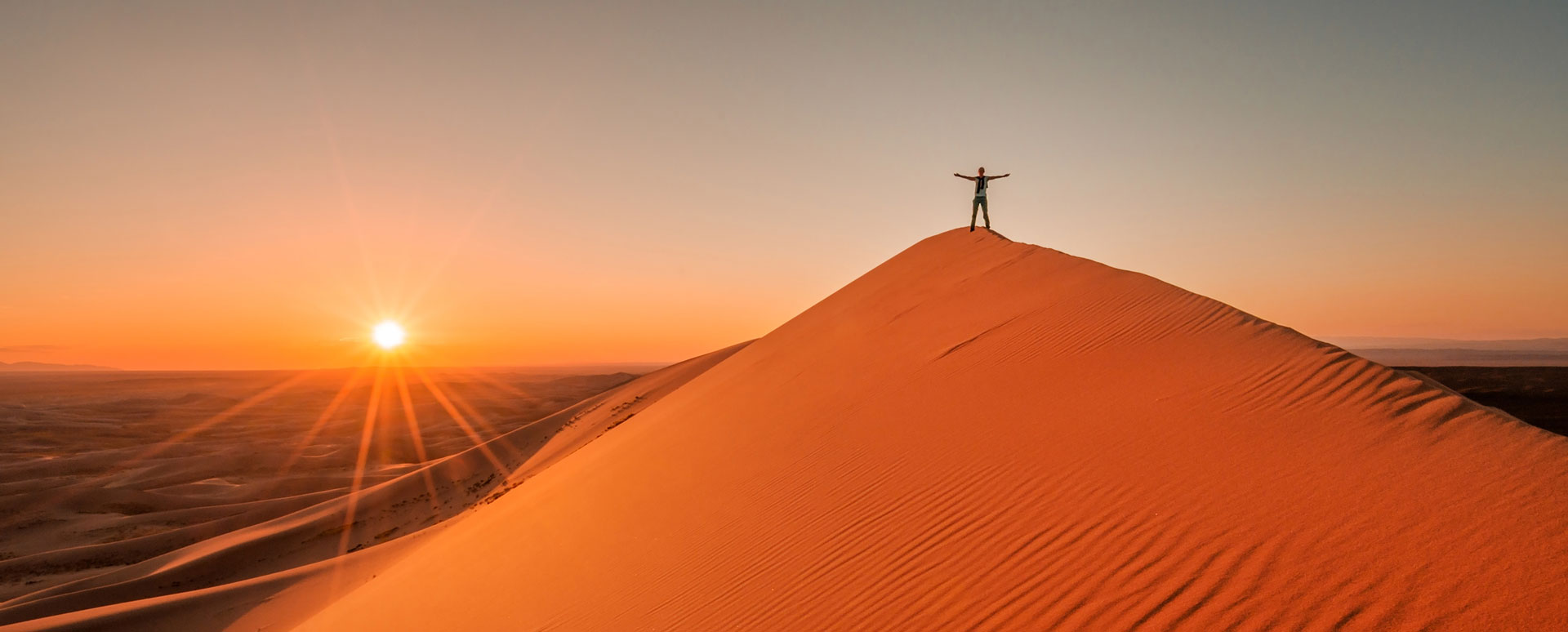 Traveller standing atop a tall sand dune with arms outstretched at sunrise in the Gobi