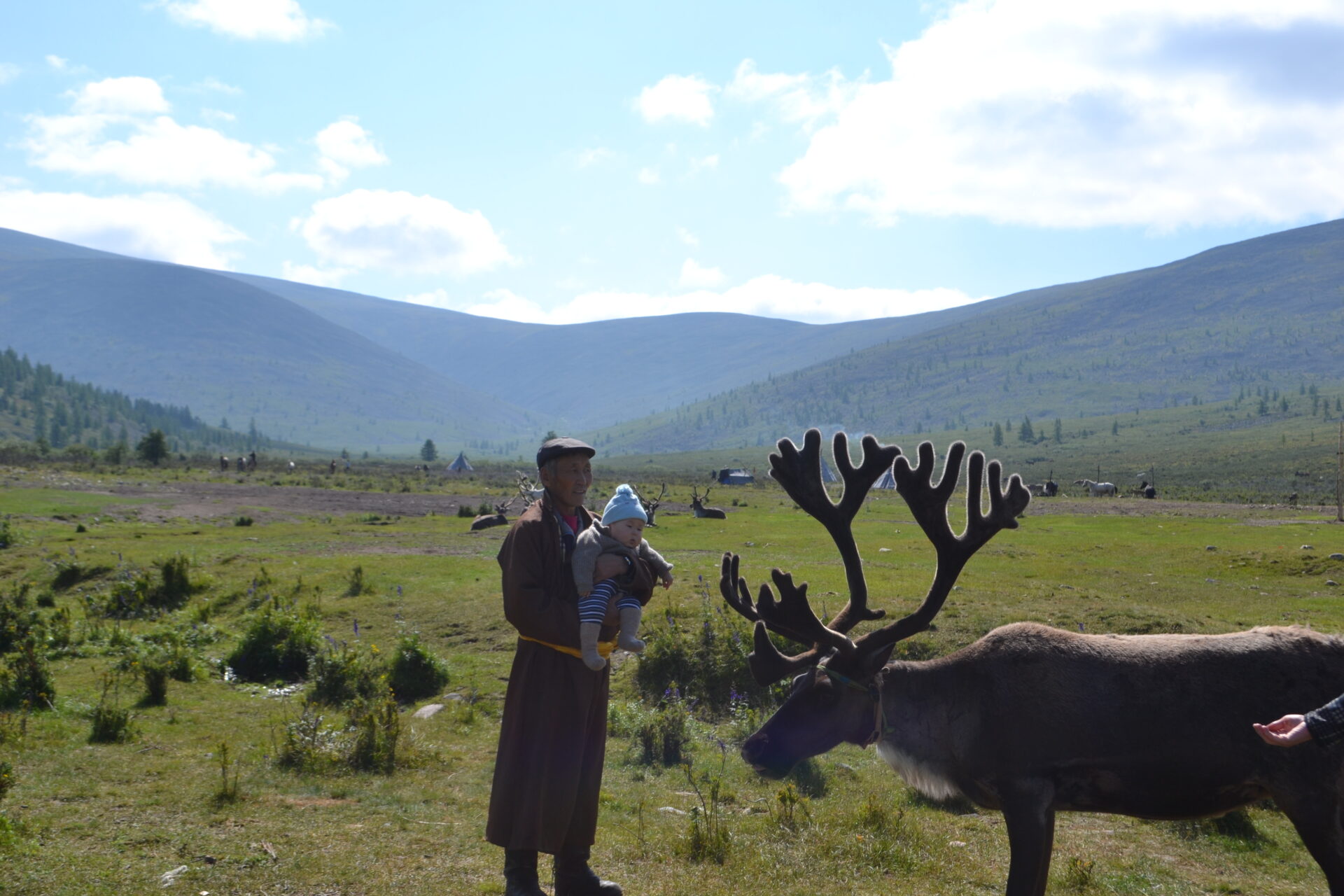 Tsaatan elder holding a baby beside a reindeer in the taiga valley
