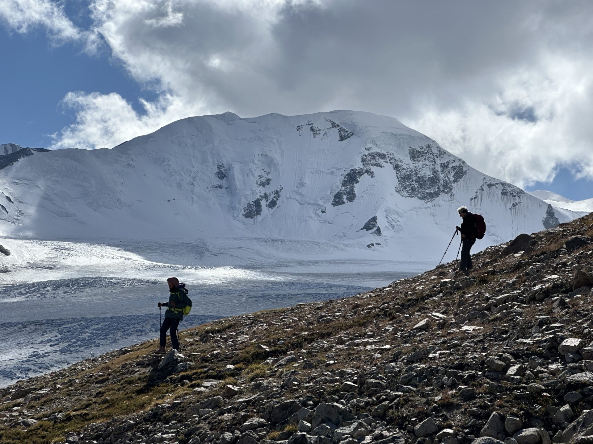 Two hikers on a rocky ridge below the snow-covered Malchin massif and its glacier